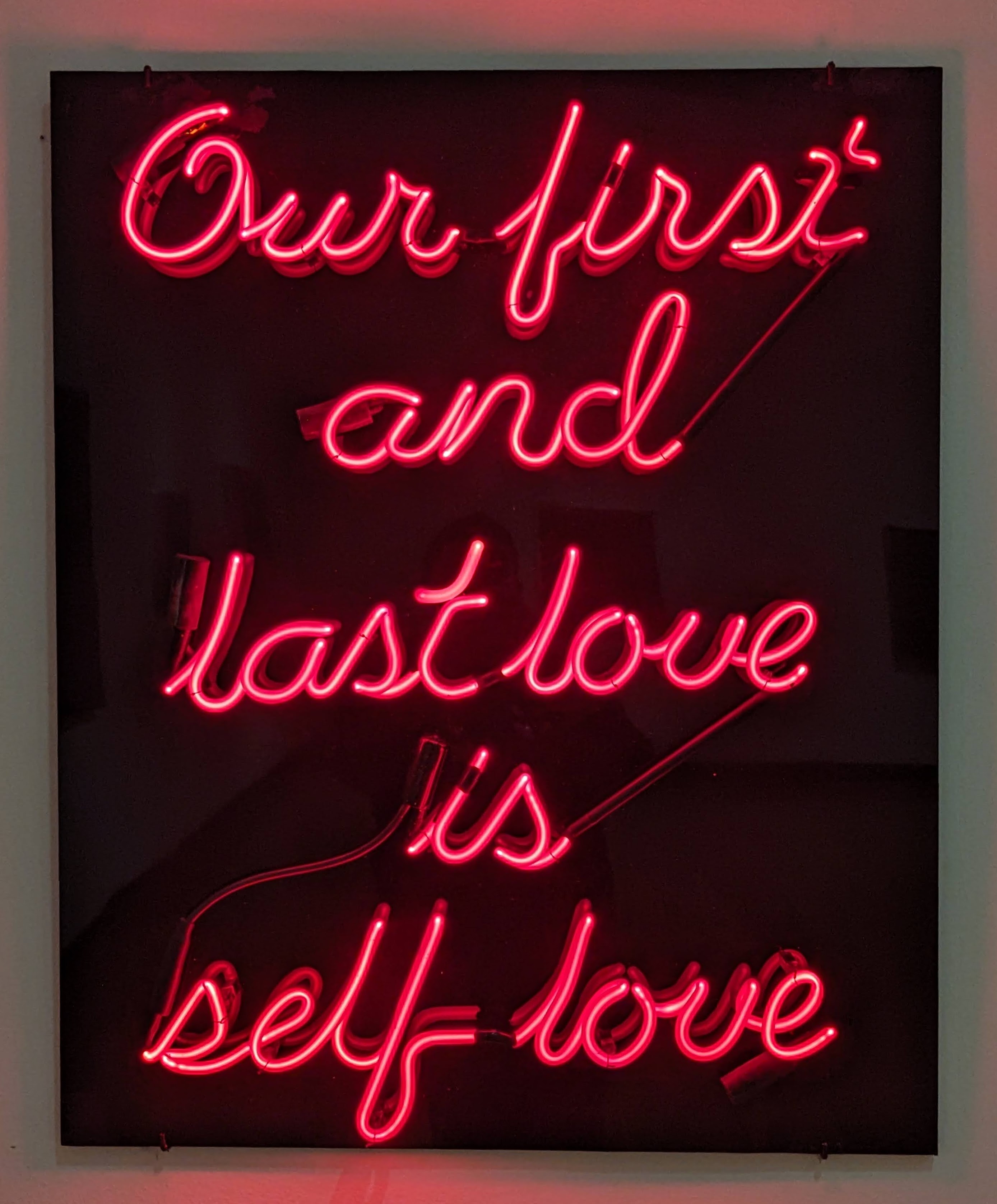 A neon sign by Black queer artist Lyle Ashton Harris reading 'Our first and last love is self love' in lit red text on a black background.